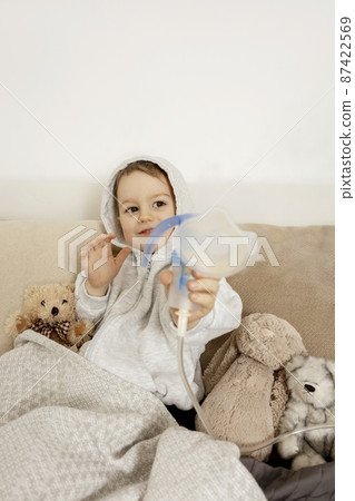 Sick little boy with inhaler for cough treatment. Unwell kid doing inhalation on his bed. Flu season. Medical procedure at home. Interior and clothes in natural earth colors. Cozy environment. Sick little boy with inhaler for cough treatment. Unwell kid doing inhalation on his bed. Flu season. Medical procedure at home. Interior and clothes in natural earth colors. Cozy environment. 87422569