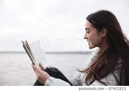 Woman reading book near river on cloudy day 87422813