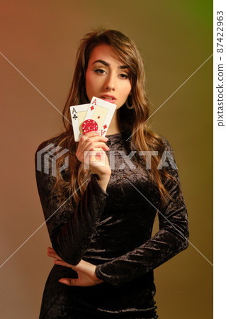 Brunette female in black dress showing two red chips and aces, posing against colorful studio background. Gambling, poker, casino. Close-up. Brunette female in black dress showing two red chips and aces, posing against colorful studio background. Gambling, poker, casino. Close-up. 87422963
