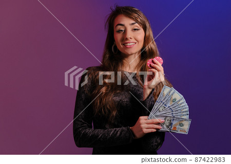 Brunette girl in black velvet dress showing two red chips and some cash, posing against coloful background. Gambling, poker, casino. Close-up. Brunette girl in black velvet dress showing two red chips and some cash, posing against coloful background. Gambling, poker, casino. Close-up. 87422983