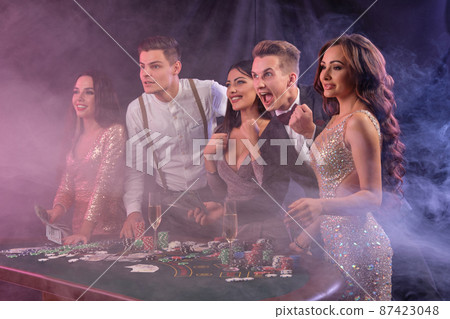 Friends playing poker at casino, at table with stacks of chips, money, cards, champagne on it. Celebrating win, excited. Black background. Close-up. 87423048