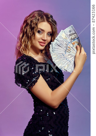 Brown-haired lady in black shiny dress is smiling, holding some cash, posing sideways on colorful background. Gambling, poker, casino. Close-up. Brown-haired lady in black shiny dress is smiling, holding some cash, posing sideways on colorful background. Gambling, poker, casino. Close-up. 87423166