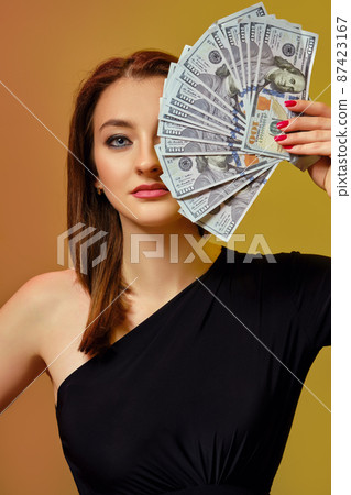 Blonde model in black dress has covered half of her face with some cash, posing against colorful background. Poker, casino. Close-up. Blonde model in black dress has covered half of her face with some cash, posing against colorful background. Poker, casino. Close-up. 87423167