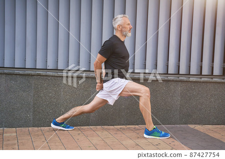 Confident sportive middle aged man in sports clothing stretching his legs, warming up and exercising outdoors Confident sportive middle aged man in sports clothing stretching his legs, warming up and exercising outdoors 87427754