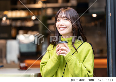 A young woman in a green sweater standing in front of a cafe with a drink 87428744