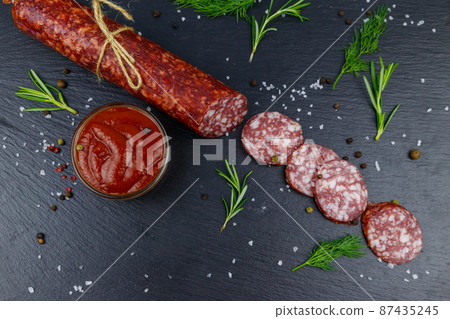 Salami sausage with ketchup and spices on black slate board. Top view Salami sausage with ketchup and spices on black slate board. Top view 87435245