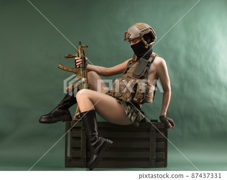 a girl soldier armed with an automatic rifle in military clothes sits on a gun case on a green background a girl soldier armed with an automatic rifle in military clothes sits on a gun case on a green background 87437331