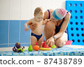Early age swimming in pool. Baby boy waiting near side, learning shapes and fruits. Happy child with coach woman in indoor swimming pool playing and having fun. Healthy and sport family with infant 87438789