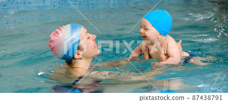Early age swimming in pool. Baby boy trained to swim in water. Happy child with trainer woman in indoor swimming pool playing and having fun. Healthy and sport family with infant, active parent 87438791
