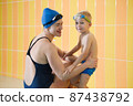 Mother instructor with toddler boy child before swimming lesson in swimming pool over yellow tile wall in goggles and hats, swimwear. Kids early physical development 87438792