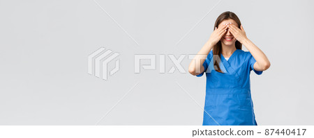 Healthcare workers, prevent virus, insurance and medicine concept. Young optimistic female nurse, doctor in scrubs, shut eyes with hands and smiling, anticipating surprise, being blindfolded 87440417