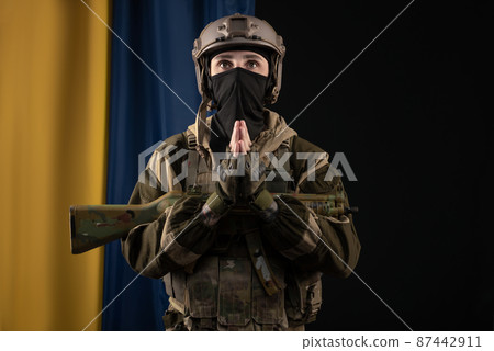 a male soldier in a military uniform and helmet with a weapon on the background of the national flag of Ukraine prays for peace 87442911