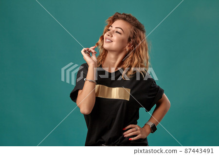Ginger girl with a curly hair, wearing black t-shirt with a golden stripe is posing standing against a blue studio background. Close-up shot. 87443491