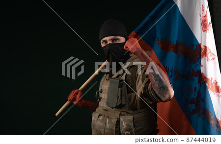 a male soldier in a military uniform with a balaclava and hands covered in blood holds a bloody flag of Russia on a black background a male soldier in a military uniform with a balaclava and hands covered in blood holds a bloody flag of Russia on a black background 87444019