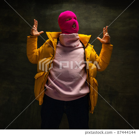 Studio shot of young anonymous man wearing pink balaclava and yellow down jacket, coat isolated on dark vintage background. Concept of safety, art, fashion Studio shot of young anonymous man wearing pink balaclava and yellow down jacket, coat isolated on dark vintage background. Concept of safety, art, fashion 87444144