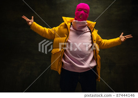 Studio shot of young anonymous man wearing pink balaclava and yellow down jacket, coat isolated on dark vintage background. Concept of safety, art, fashion Studio shot of young anonymous man wearing pink balaclava and yellow down jacket, coat isolated on dark vintage background. Concept of safety, art, fashion 87444146