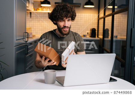 Portrait of happy surprised young curly man opening gift box with present during video call on laptop computer sitting at table in kitchen with modern interior. Concept of remote celebration. Portrait of happy surprised young curly man opening gift box with present during video call on laptop computer sitting at table in kitchen with modern interior. Concept of remote celebration. 87449868