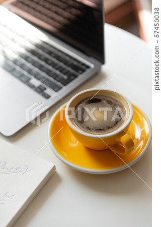 Yellow cup of coffee on workspace on table near laptop.  87450038