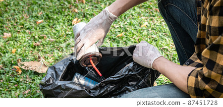 Volunteer man in gloves sitting to picking up coffee cup into plastic black bag for cleaning the park Volunteer man in gloves sitting to picking up coffee cup into plastic black bag for cleaning the park 87451728