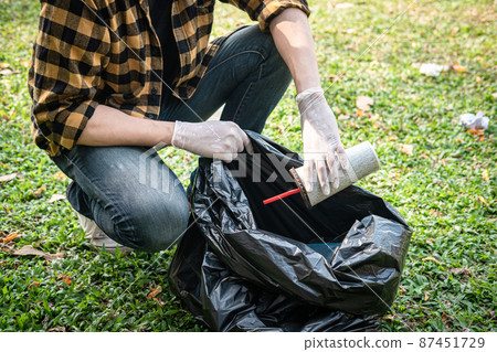Volunteer man in gloves sitting to picking up coffee cup into plastic black bag for cleaning the park Volunteer man in gloves sitting to picking up coffee cup into plastic black bag for cleaning the park 87451729