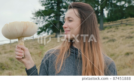 Close-up, happy woman holds red amanita in hand, back of fir forest in autumn day. Huge orange toxic fly agaric toadstool in girl's hand. Beginning mushroom picking season. Deadly red fungus in fall Close-up, happy woman holds red amanita in hand, back of fir forest in autumn day. Huge orange toxic fly agaric toadstool in girl's hand. Beginning mushroom picking season. Deadly red fungus in fall 87451888