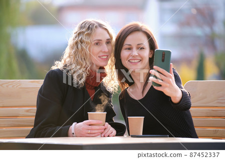 Two happy women making selfie with mobile phone sitting at city street cafe. Female friends enjoying time together outdoors Two happy women making selfie with mobile phone sitting at city street cafe. Female friends enjoying time together outdoors 87452337