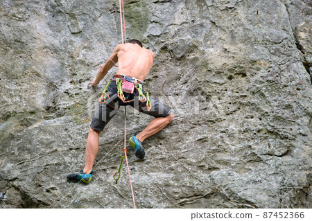 Strong male climber climbing steep wall of rocky mountain. Sportsman overcoming difficult route. Engaging in extreme sports hobby concept. 87452366