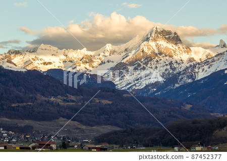Small town at foot of magnificent Alps mountain range, Switzerland. Quiet life on background of fantastic peaks covered with shiny snow and dark green forest under blue sky with puffy white clouds. Small town at foot of magnificent Alps mountain range, Switzerland. Quiet life on background of fantastic peaks covered with shiny snow and dark green forest under blue sky with puffy white clouds. 87452377