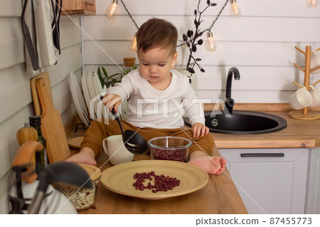 A little brunet boy, sits on the kitchen table, plays with cutlery and beans, 87455773