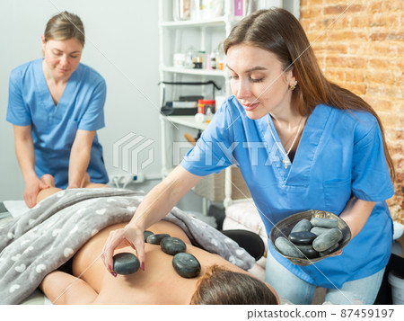 Two masseuses massage the legs and perform a stone therapy procedure Two masseuses massage the legs and perform a stone therapy procedure 87459197
