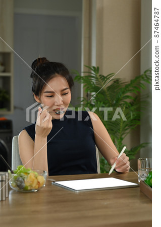 Healthy young woman using digital tablet and eating fresh vegetable salad at home kitchen. 87464787