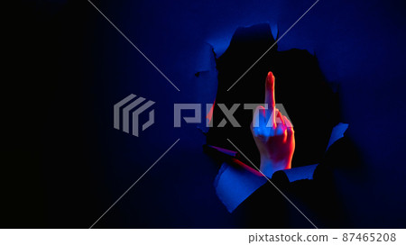 Fuck you. Provocative gesture. Red blue neon light female hand showing middle finger obscene sign inside breakthrough paper hole isolated on dark night copy space banner. 87465208