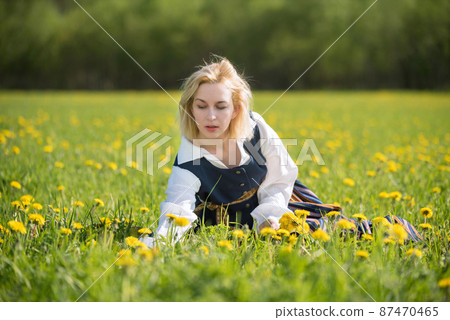 Young woman in national clothes wearing yellow dandelion wreath in spring field. Springtime Young woman in national clothes wearing yellow dandelion wreath in spring field. Springtime 87470465