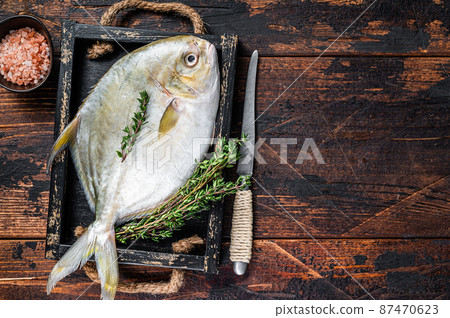 Raw fish butterfish or pompano with herbs in a wooden tray. Dark wooden background. Top view. Copy space Raw fish butterfish or pompano with herbs in a wooden tray. Dark wooden background. Top view. Copy space 87470623