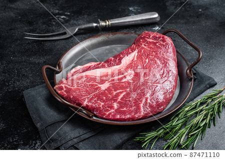 Raw top sirloin beef meat steak in a steel tray with rosemary. Black background. Top view 87471110