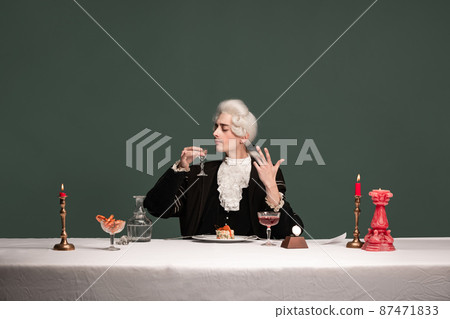 Portrait of young elegant man in peruke and vintage jacket sitting at table isolated on dark green background. Retro style, comparison of eras concept. Portrait of young elegant man in peruke and vintage jacket sitting at table isolated on dark green background. Retro style, comparison of eras concept. 87471833