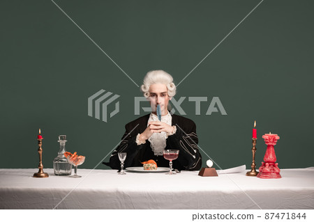 Portrait of young elegant man in peruke and vintage jacket sitting at table isolated on dark green background. Retro style, comparison of eras concept. Portrait of young elegant man in peruke and vintage jacket sitting at table isolated on dark green background. Retro style, comparison of eras concept. 87471844