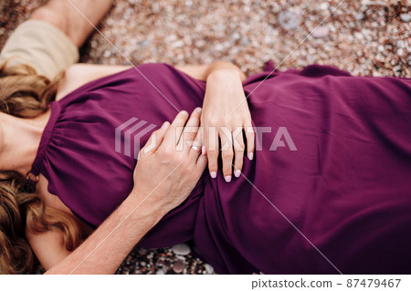 Bride in a burgundy dress lies on a pebble with her hands folded on her stomach. Close-up Bride in a burgundy dress lies on a pebble with her hands folded on her stomach. Close-up 87479467