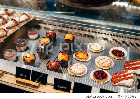 Various cakes are on plates in a display case with price tags 87479562