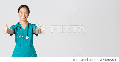 Covid-19, healthcare workers, pandemic concept. Smiling pretty asian female doctor, nurse in scrubs ensure everything good, showing thumbs-up in approval, agree or like something, white background Covid-19, healthcare workers, pandemic concept. Smiling pretty asian female doctor, nurse in scrubs ensure everything good, showing thumbs-up in approval, agree or like something, white background 87480904