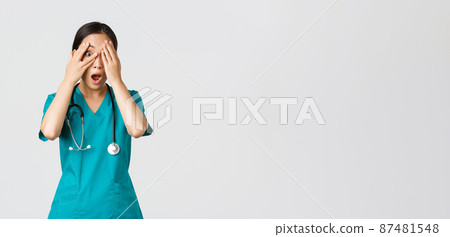 Covid-19, healthcare workers and preventing virus concept. Scared and shocked asian female nurse, doctor in scrubs witness something embarrassing or scary, cover eyes but peek through fingers 87481548
