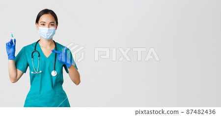 Covid-19, coronavirus disease, healthcare workers concept. Confident friendly-looking asian female doctor, physician in medical mask and scrubs pointing finger at syringe with vaccine 87482436
