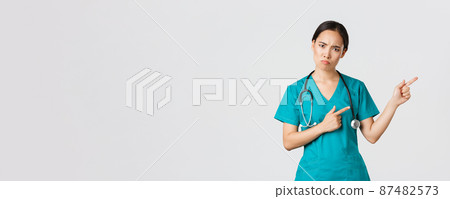 Covid-19, healthcare workers, pandemic concept. Disappointed and angry asian female therapist, nurse in scrubs looking upset at camera, sulking and pointing upper right corner, wait explanation 87482573