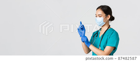 Covid-19, coronavirus disease, healthcare workers concept. Professional smiling asian female nurse, physician in scrubs and medical mask put on rubber gloves for checkup, patient examination 87482587
