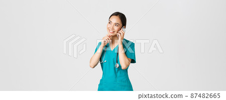 Covid-19, healthcare workers and preventing virus concept. Portrait of smiling asian female doctor, intern in scrubs talking on phone and looking thoughtful, thinking or making choice 87482665