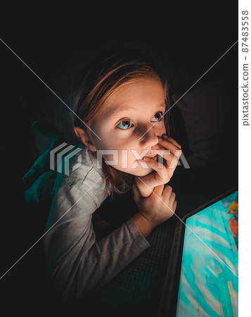Little caucasian girl looking at a tablet in the evening in the dark on the bed Little caucasian girl looking at a tablet in the evening in the dark on the bed 87483558