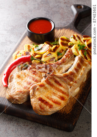 Pork chops with grilled potatoes, sauce and chili peppers close-up on a wooden board. vertical Pork chops with grilled potatoes, sauce and chili peppers close-up on a wooden board. vertical 87483601