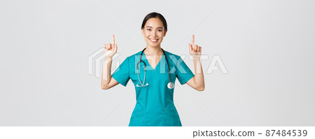 Covid-19, healthcare workers, pandemic concept. Happy smiling asian female doctor, nurse in scrubs showing advertisement, making announcement. Physician pointing fingers up at banner Covid-19, healthcare workers, pandemic concept. Happy smiling asian female doctor, nurse in scrubs showing advertisement, making announcement. Physician pointing fingers up at banner 87484539