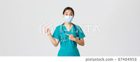 Covid-19, coronavirus disease, healthcare workers concept. Surprised and interested asian female doctor, physician in medical mask and scrubs pointing fingers upper left corner, white background 87484958