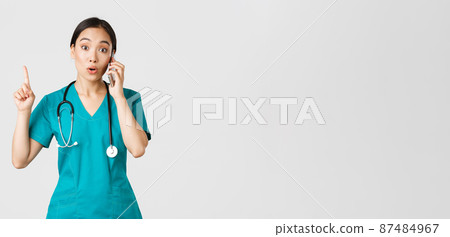 Covid-19, healthcare workers and preventing virus concept. Portrait of beautiful asian female doctor, nurse in scrubs talking on phone and raise finger as have great idea, remember, suggest plan 87484967